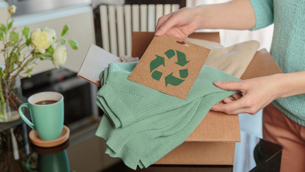 Fast Fashion vs Sustainable Fashion: A Smarter, More Sustainable Way to Shop 2 A person unpacks a cardboard box containing a green sweater with a recycling symbol tag, highlighting the contrast between fast fashion vs sustainable fashion. A cup of coffee and white flowers are on the table in the background.