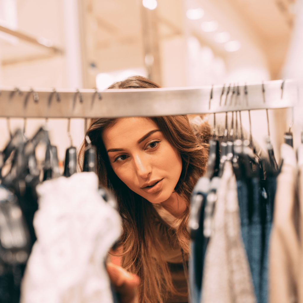Greenwashing: How to Spot a Truly Sustainable Brand 1 A person with long, wavy brown hair is browsing through a clothing rack in a store. They are holding a white garment in one hand while looking through various clothes, including jeans and sweaters, hanging on the rack. The background shows a well-lit retail space with blurred shelves and lights.