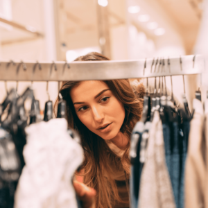 A person with long, wavy brown hair is browsing through a clothing rack in a store. They are holding a white garment in one hand while looking through various clothes, including jeans and sweaters, hanging on the rack. The background shows a well-lit retail space with blurred shelves and lights.