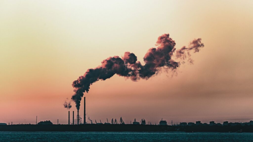 Fast Fashion vs Sustainable Fashion: A Smarter, More Sustainable Way to Shop 1 Thick, dark smoke billows from factory chimneys into a sunset sky, silhouetting industrial structures by the water, illustrating the environmental impact of fast fashion.